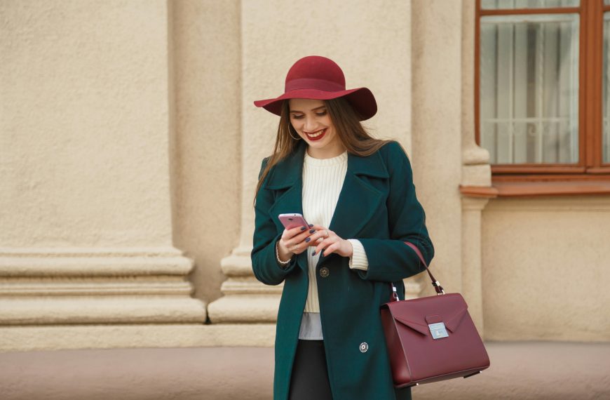 woman wearing a maroon hat and a dark teal coat over a white top, carrying a maroon bag